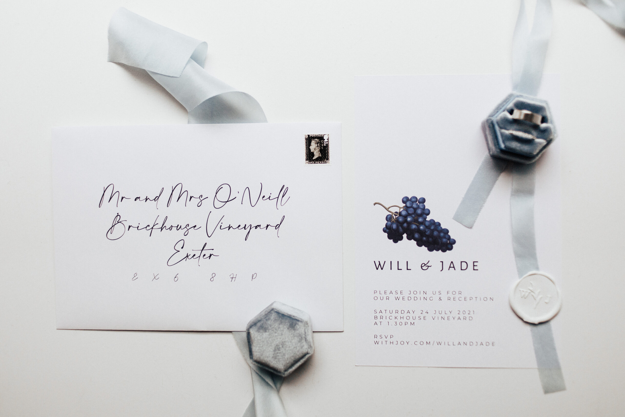 A wedding flat lay of a black and white bespoke wedding invitation and envelope. The invitation reads "Will and Jade" and has blue grapes. The envelope has modern calligraphy and a black stamp. The flat lay also includes blue and white details such as a blue ring box with silver wedding rings, blue ribbon, and a white personalised initials wax seal.