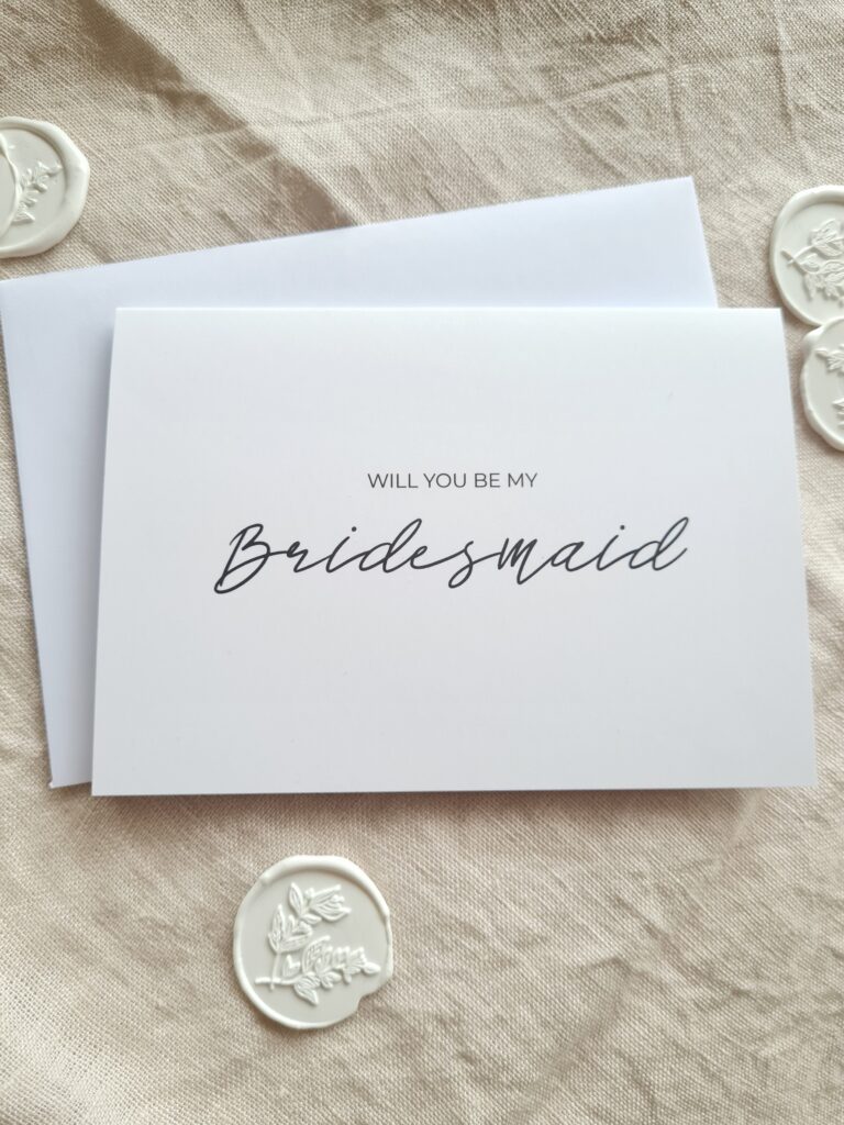A greeting card with elegant calligraphy that reads "Will you be my bridesmaid". The card has a simple and modern design, with a white background and black text.