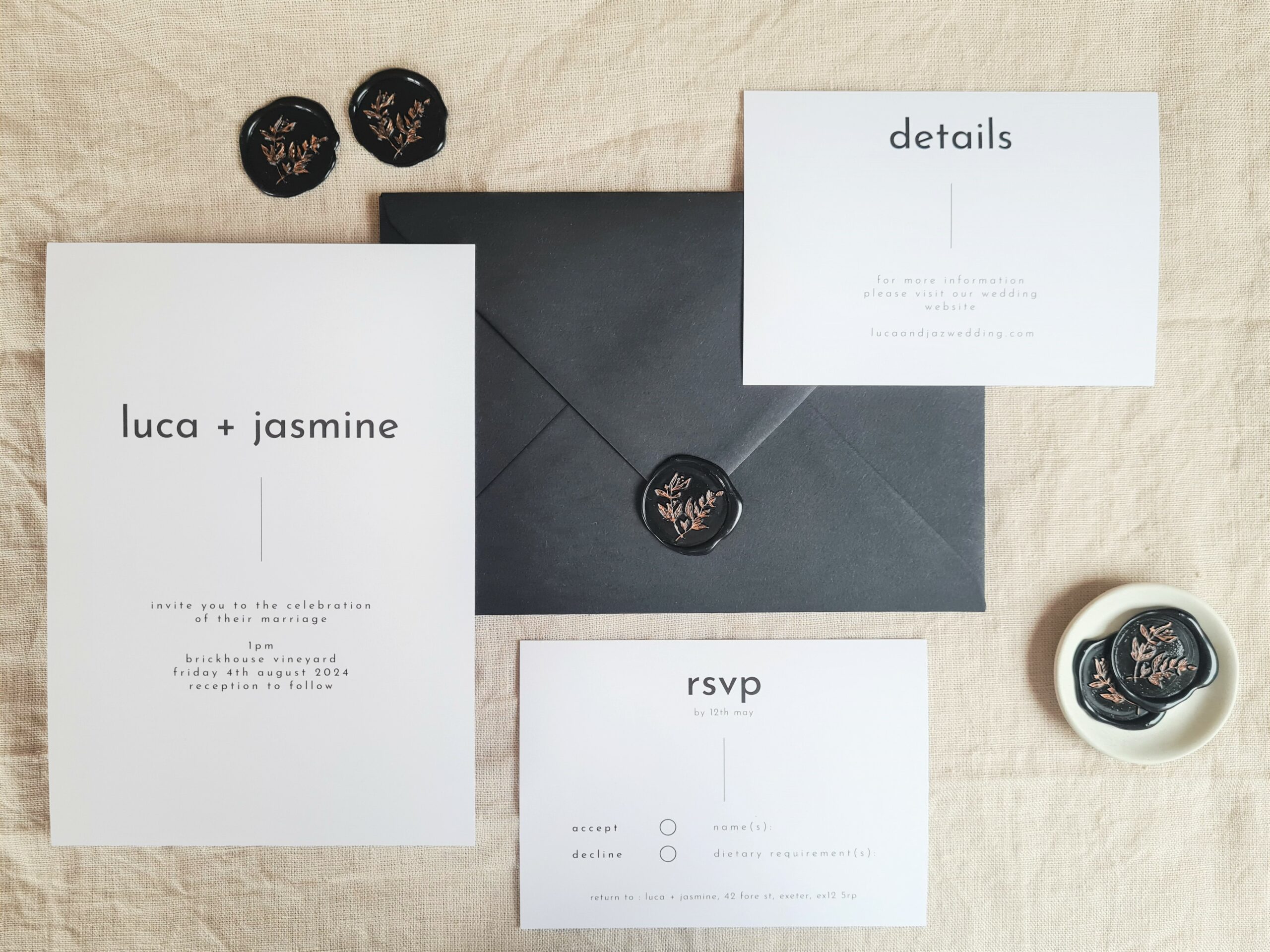 A black and white invitation suite flat lay. There is an RSVP card, invite and Details card. They are set against a beige linen background. Black wax seals, a mini white plate and a black envelope is arranged around it.