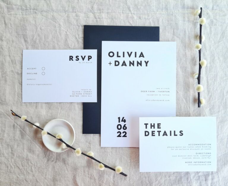 A black and white invitation suite with the words "Olivia and Danny" written on it. There is an RSVP card, invite and Details card. They are set against a beige linen background. Modern dried flowers are arranged around it and a white mini plate and a black envelope.