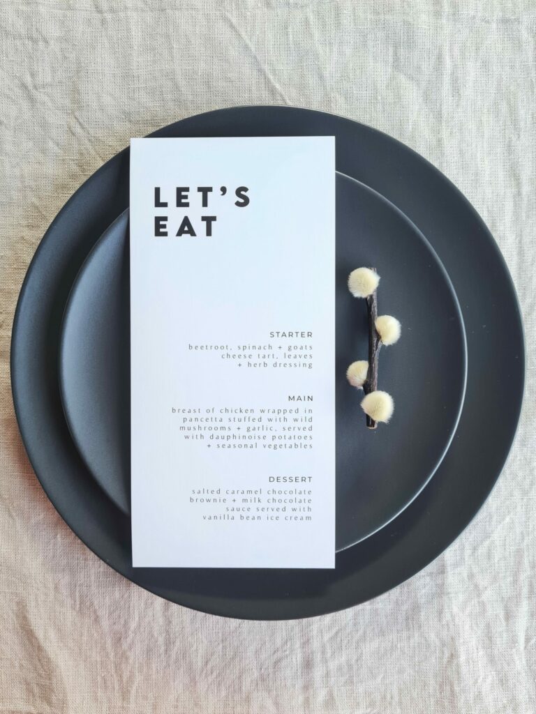 A black and white wedding menu reading "Let's eat" is elegantly placed on a black plate, with bold font and intricate floral details adding a touch of sophistication. The setting is arranged on a beige linen background, creating a neutral yet refined aesthetic.