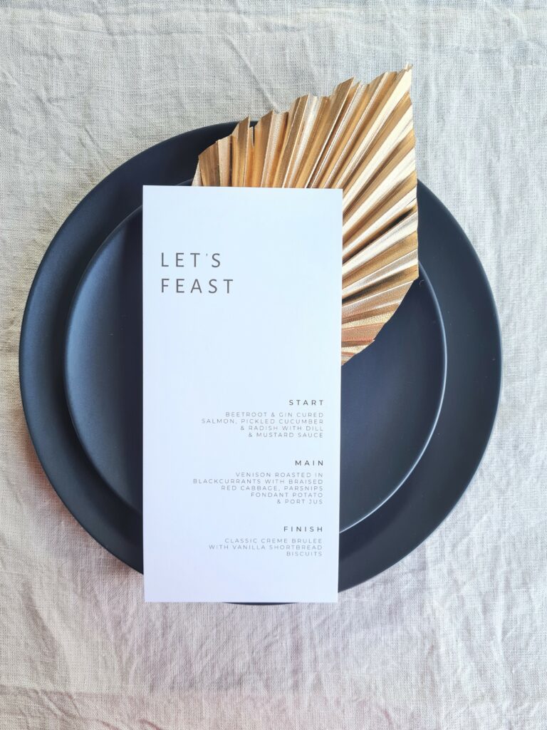 A black and white wedding menu with the text "Let's Feast" is placed on a black plate. The plate is set on a beige linen background and is accompanied by a gold leaf fan. The overall aesthetic is elegant and sophisticated.