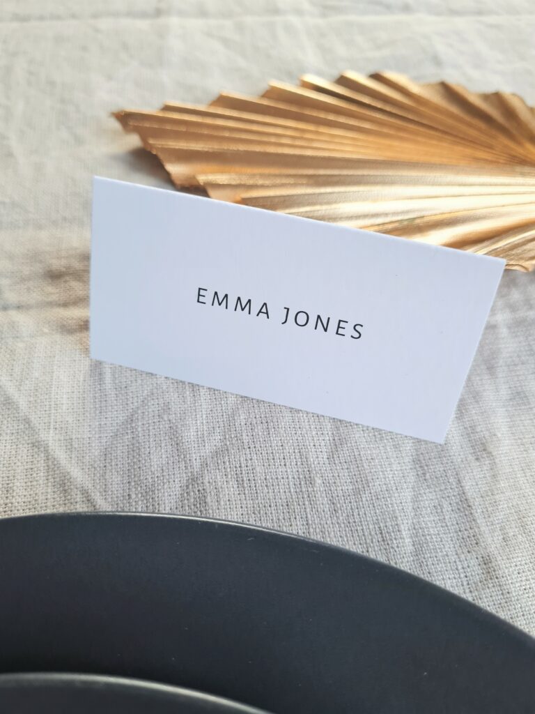 A black and white tent-fold place card is elegantly placed next to a black plate with a gold leaf fan accent, all set against a beige linen background. The combination of the black and gold elements exudes a sense of modern elegance
