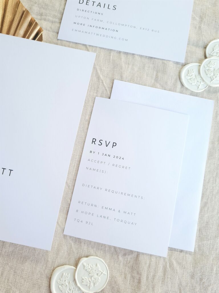 A black and white details card, invite and RSVP card are placed on a beige linen background and is accompanied by a gold leaf fan and white wax seals with botanical detailing. The overall aesthetic is elegant and sophisticated.