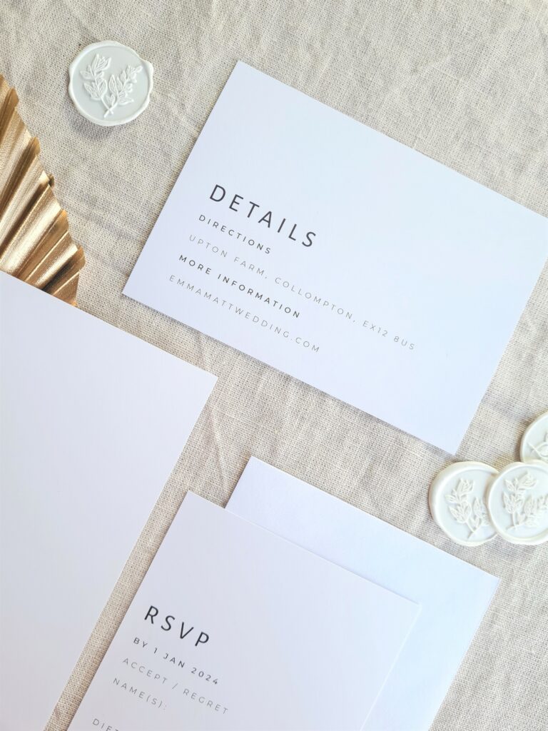A black and white details card and RSVP card are placed on a beige linen background and is accompanied by a gold leaf fan and white wax seals with botanical detailing. The overall aesthetic is elegant and sophisticated.