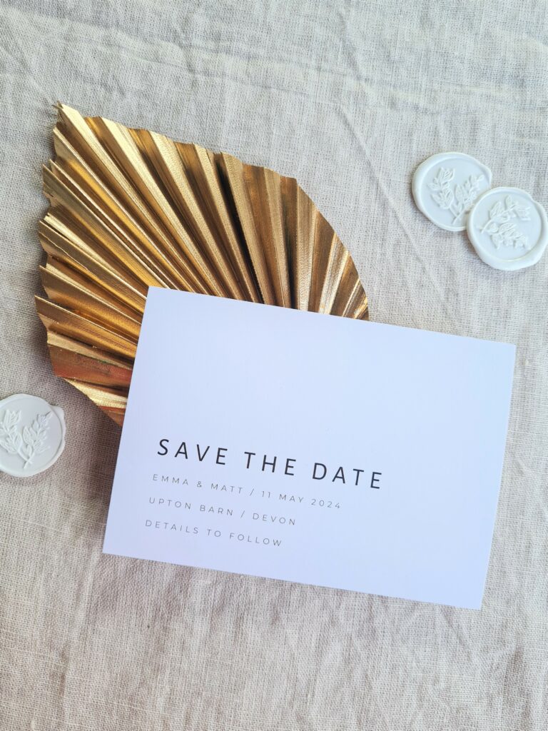 A black and white save the date card is placed on a beige linen background and is accompanied by a gold leaf fan and white wax seals with botanical detailing. The overall aesthetic is elegant and sophisticated.