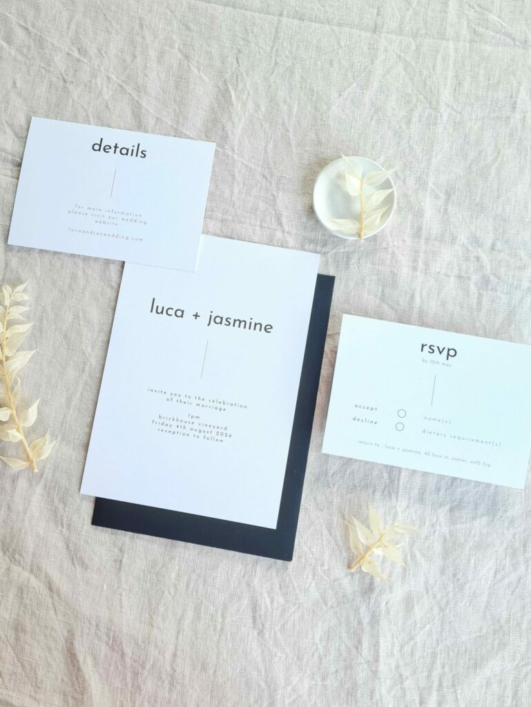 A black and white invitation suite flat lay. There is an RSVP card, invite and Details card. They are set against a beige linen background. Modern dried flowers are arranged around it and a white mini plate and a black envelope.