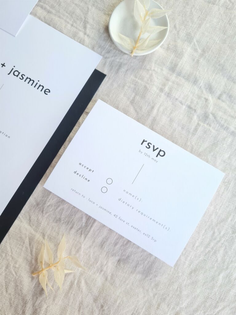 A black and white wedding invite and rsvp card is set on a beige linen background and is accompanied by a white floral elements and mini plate and a black envelope. The overall aesthetic is elegant and sophisticated.