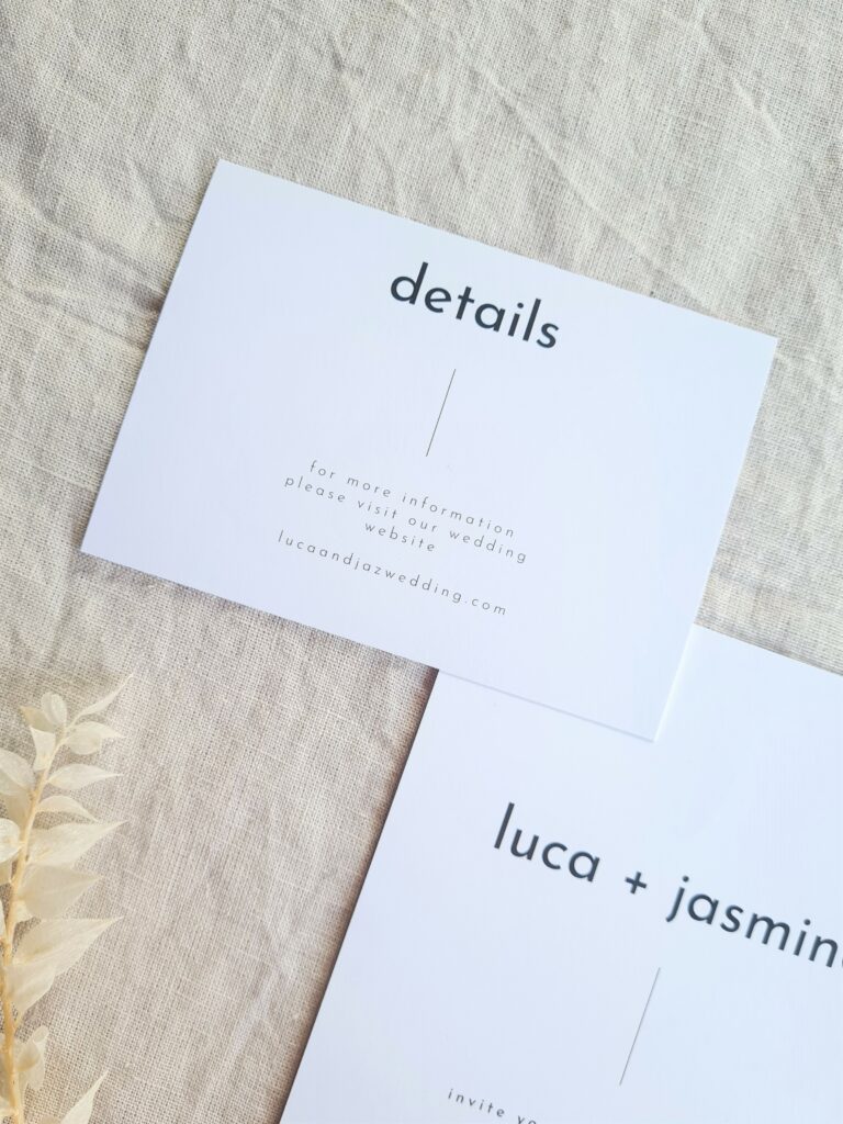 A black and white wedding invite and details card is set on a beige linen background and is accompanied by a white floral elements. The overall aesthetic is elegant and sophisticated.