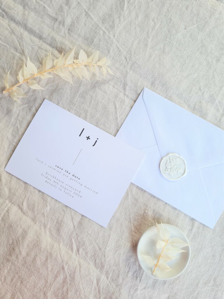 A black and white save the date card is set on a beige linen background and is accompanied by a white floral elements and mini plate and a white envelope with wax seal. The overall aesthetic is elegant and sophisticated.