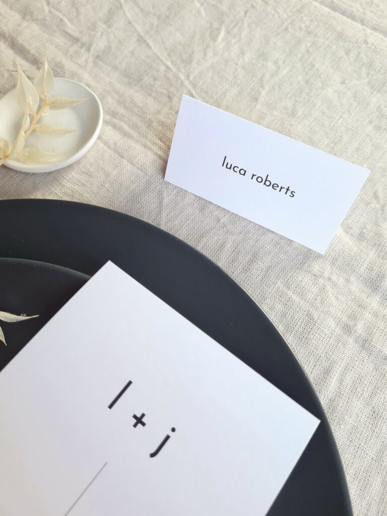A black and white tent-fold place card is elegantly placed next to a black plate and a black and white wedding menu on a beige linen background. The table is adorned with delicate white floral arrangements, adding a touch of sophistication to the overall aesthetic.