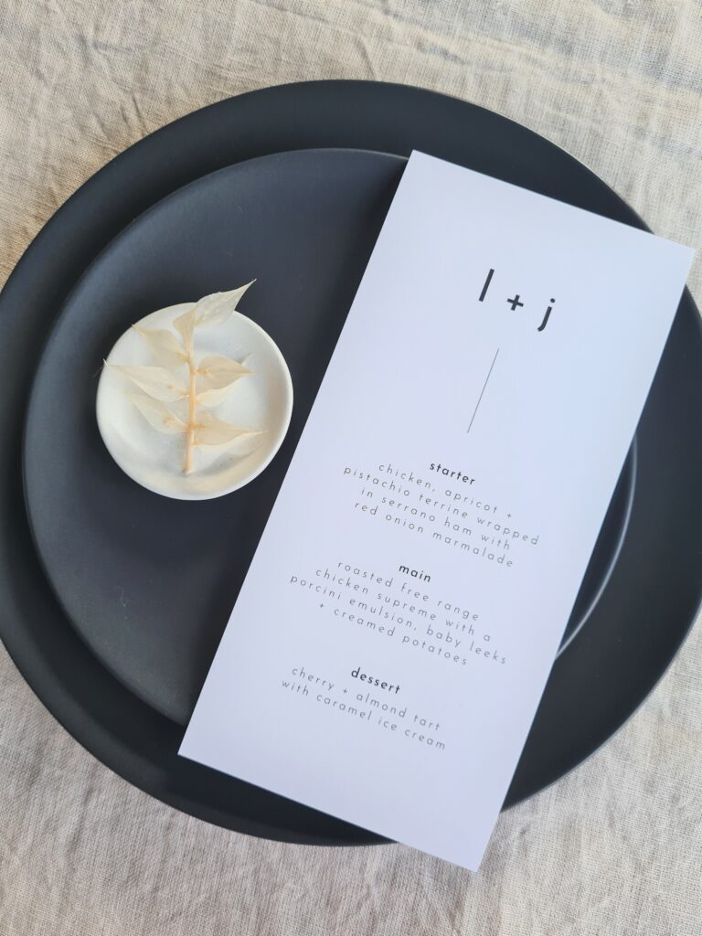 A black and white wedding menu is displayed on a sleek black plate with white details, set against a beige linen background. The table is elegantly adorned with delicate white floral arrangements, adding a touch of sophistication and romance to the overall aesthetic.