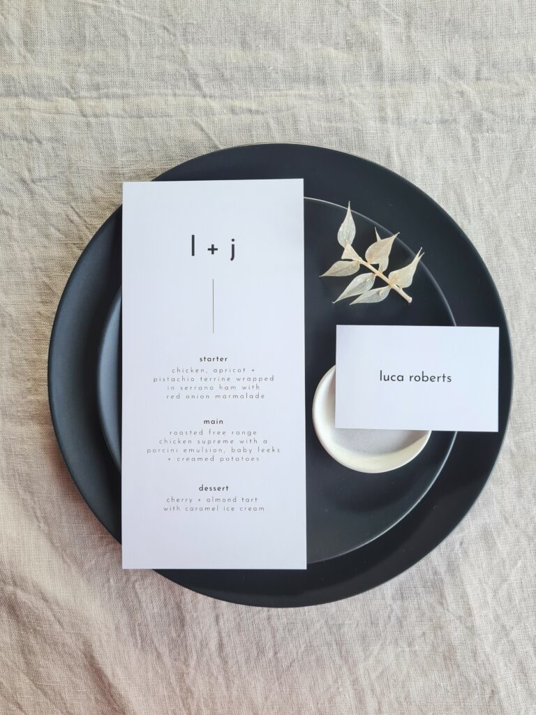 A black and white wedding menu and place card is displayed on a sleek black plate with white details, set against a beige linen background. The table is elegantly adorned with delicate white floral arrangements, adding a touch of sophistication and romance to the overall aesthetic.