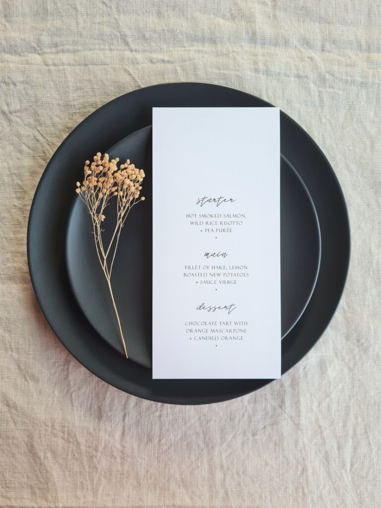 A black and white wedding menu is placed on a black plate. The plate is set on a beige linen background and is accompanied by natural dried floral elements. The overall aesthetic is elegant, modern and sophisticated.