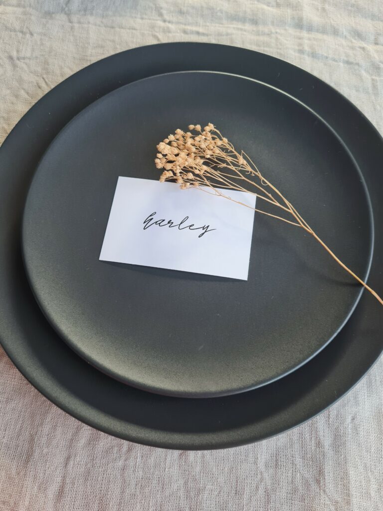 A black and white contemporary place card is elegantly placed on a black plate next to a natural dried flower The minimalistic design of the place card perfectly complements the bold font and sleek design of the menu. The entire setting is captured against a beautiful beige linen background, which adds a touch of warmth to the monochrome palette.