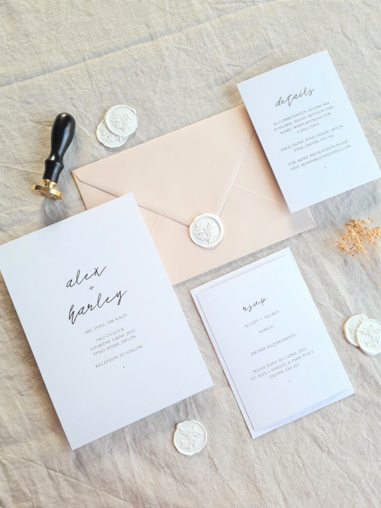 This is a flat lay image of a black and white modern wedding invitation suite set on a beige linen background with various dried flowers scattered around it. The suite includes an invitation, an RSVP card, and a details card. The save the date card is a simple black and white design, while the invitation features bold black text on a white background, with a black and gold wax stamp and white wax seals.