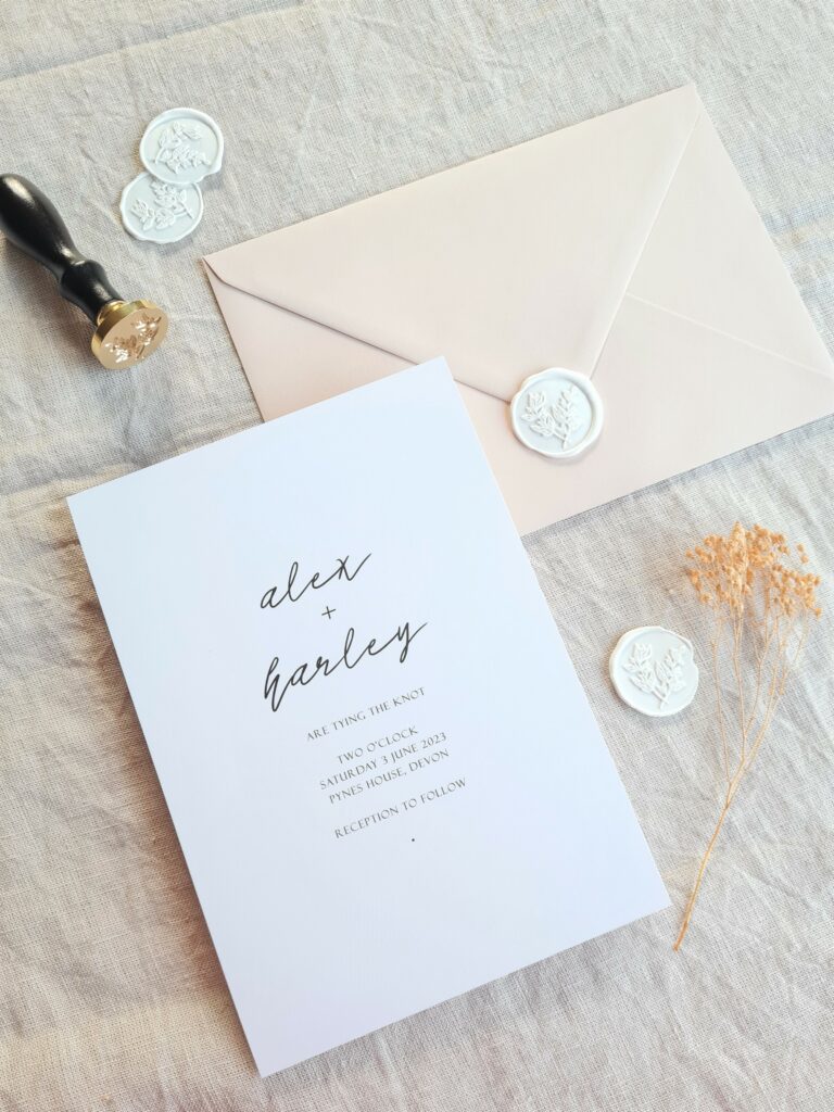 A black and white wedding invitation is placed on a beige linen background and is accompanied by natural dried floral elements, white botanical wax seals and a beige envelope. The overall aesthetic is elegant, modern and sophisticated.