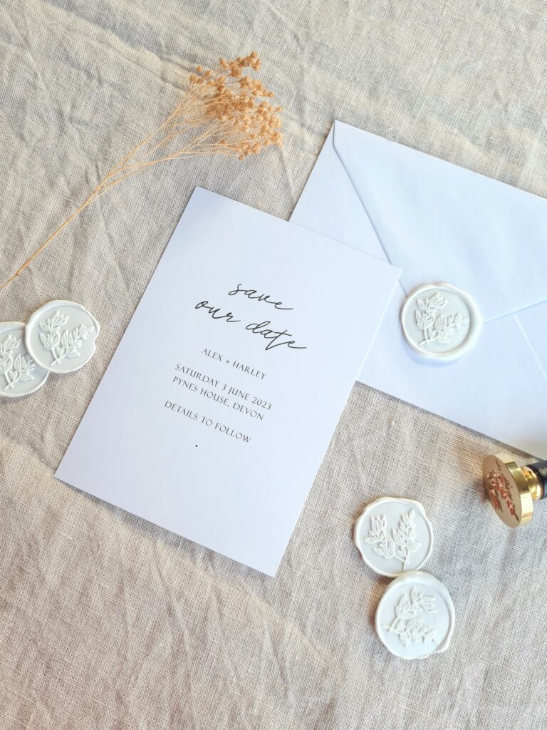 A black and white wedding save the date is placed on a beige linen background and is accompanied by natural dried floral elements, white botanical wax seals and a white envelope. The overall aesthetic is elegant, modern and sophisticated.