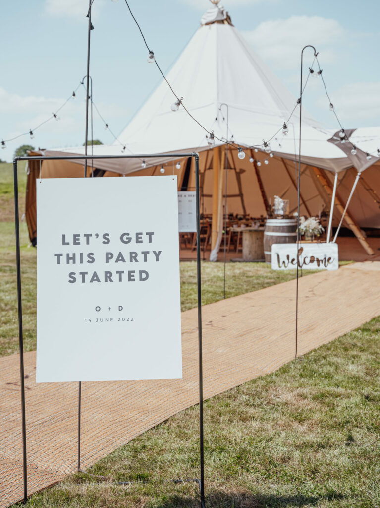A black and white wedding welcome sign reading "Let's get this party started" displayed in front of a natural coloured tipi at Deer Farm Tipi Weddings near Exeter. The sign features clear, easy-to-read text and elegant design. Natural, beige, black and white décor elements including wooden barrel, flowers, and lighting are arranged around the sign, adding a modern touch to the overall look.