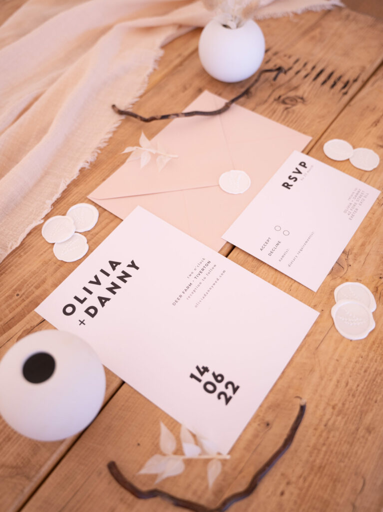 A flat lay of black and white wedding stationery items including an invitation card, RSVP card, wax seals, and a beige envelope. The items are arranged in a flat lay style on a wooden table with linen and floral details.