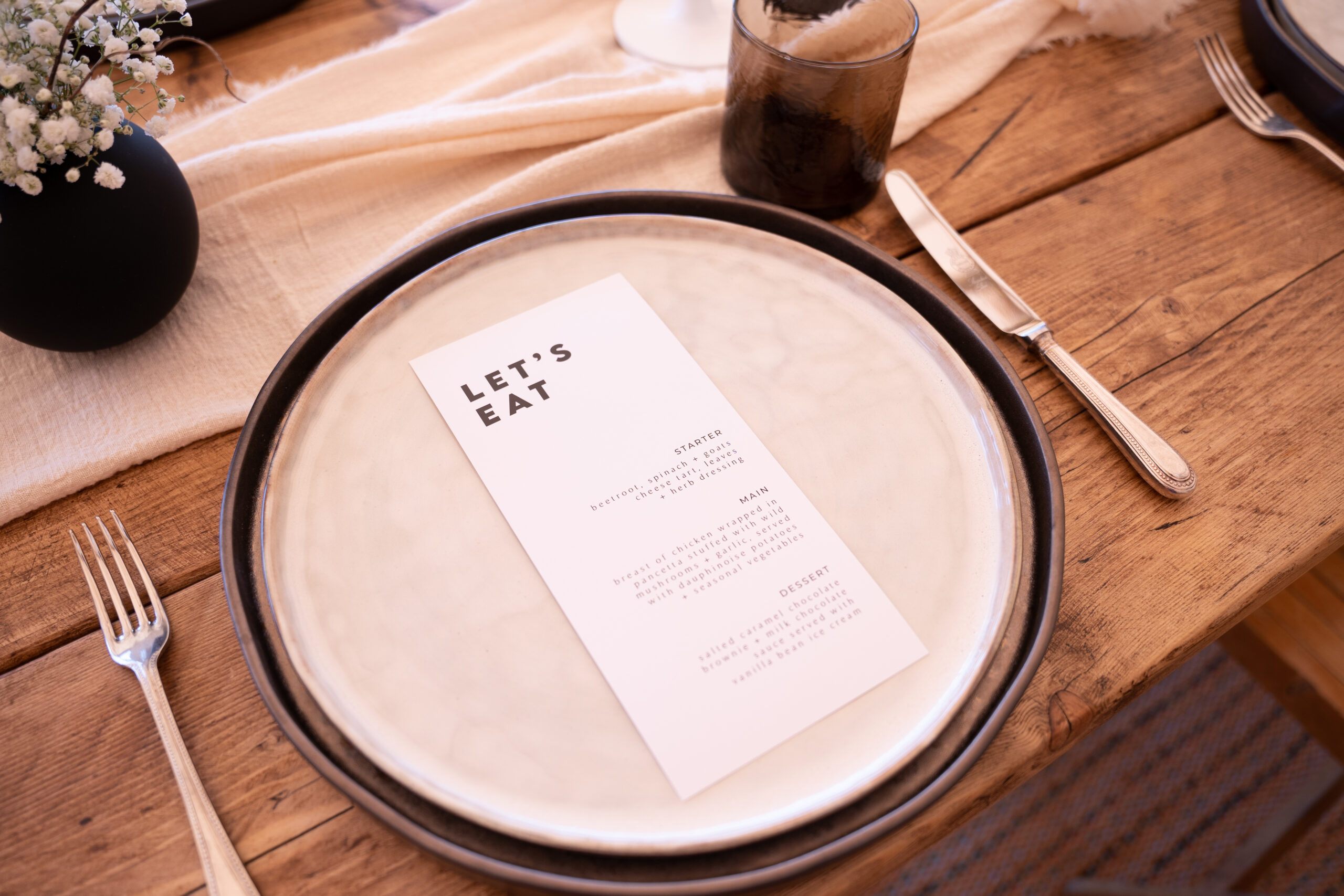 A black and white wedding menu tables cape with beige décor displayed on a table inside a tipi at Deer Farm Tipi Weddings near Exeter. The wooden table features a beige tablecloth, modern black and grey plates with a black and white floral accents.