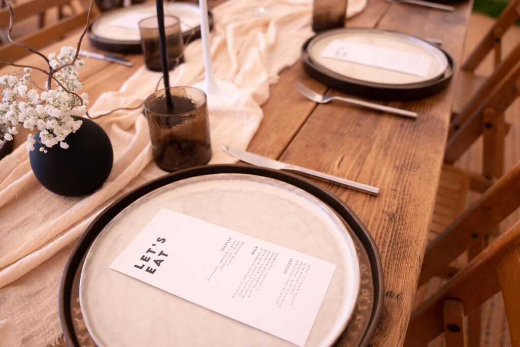 A black and white wedding menu tables cape displayed on a plate with black, white, and beige décor at Deer Farm Tipi Weddings near Exeter. The tables cape is styled with elegant and rustic elements, including a ethically made charger plate, white and beige linen, and black and white floral accents.