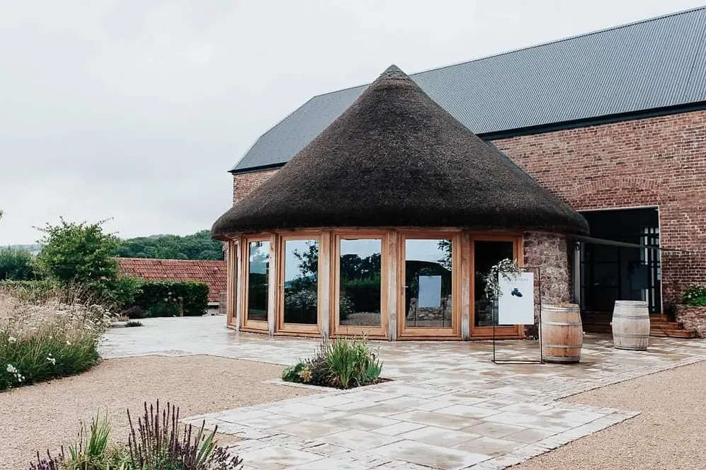 The image features the beautiful grounds of Brickhouse Vineyard located near Exeter, Devon. The picture showcases the thatched roundhouse, a red brick barn, and the surrounding vineyard. The photo also features a black frame sign with a bespoke hanging welcome sign and wine barrels in the foreground.