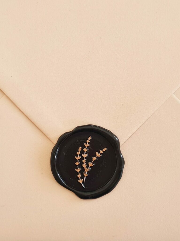 A beige envelope sealed with a black wax seal featuring bronze hand-painted foliage detailing. The seal has an intricate and detailed design with a combination of black and bronze colours that contrast well against the beige background of the envelope.