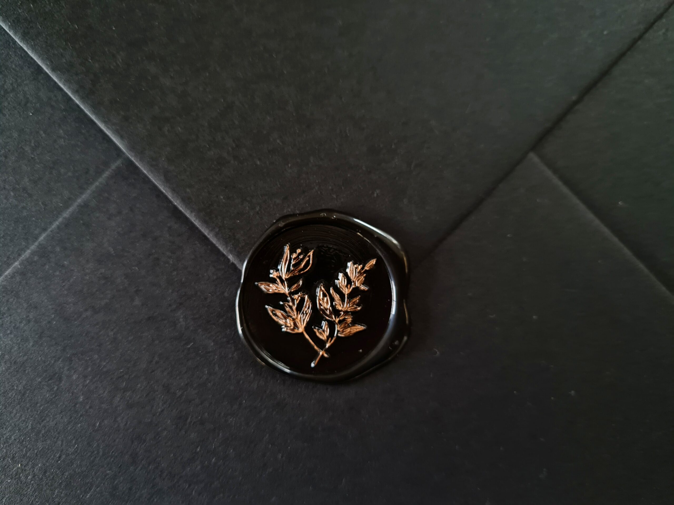 A black envelope with a black wax seal on the flap featuring bronze hand-painted leaf detailing.