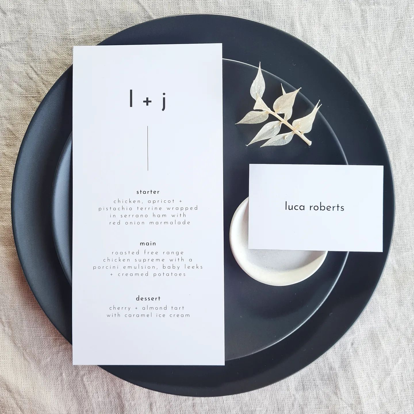 A black and white wedding menu with the text "l +j" and a place card are placed on a black plate. The plate is set on a beige linen background and is accompanied by a white floral elements and small plate. The overall aesthetic is elegant and sophisticated.
