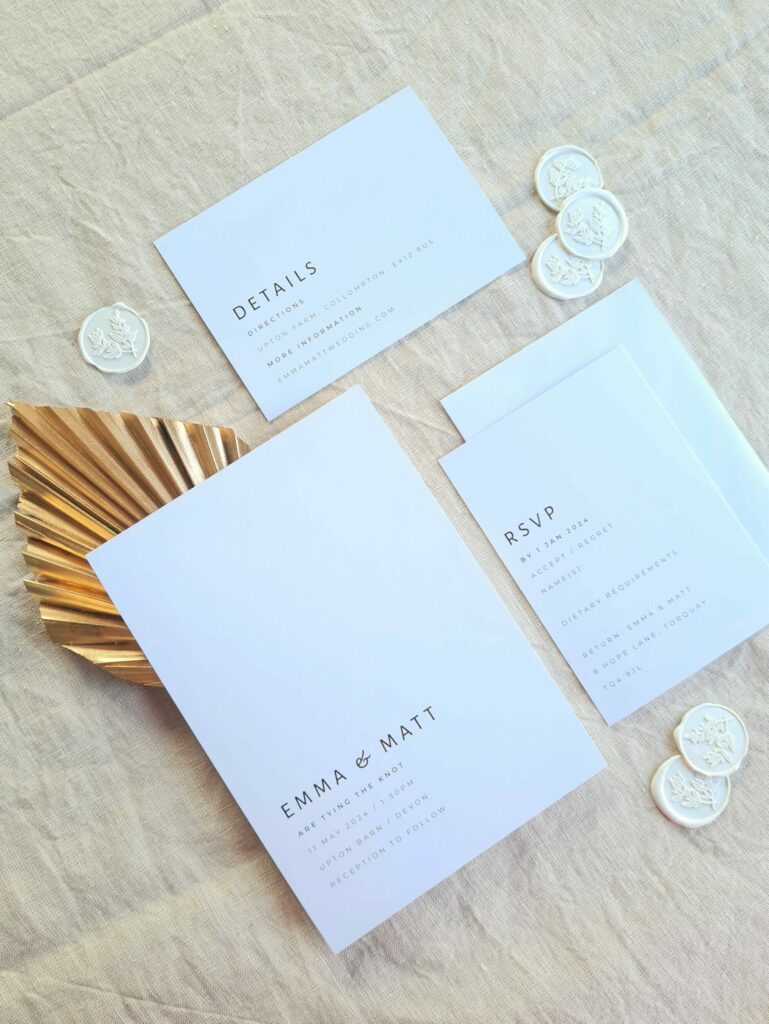 A black and white wedding invitation, RSVP card, and details card with white wax seals and a gold fan arranged on a beige linen background.