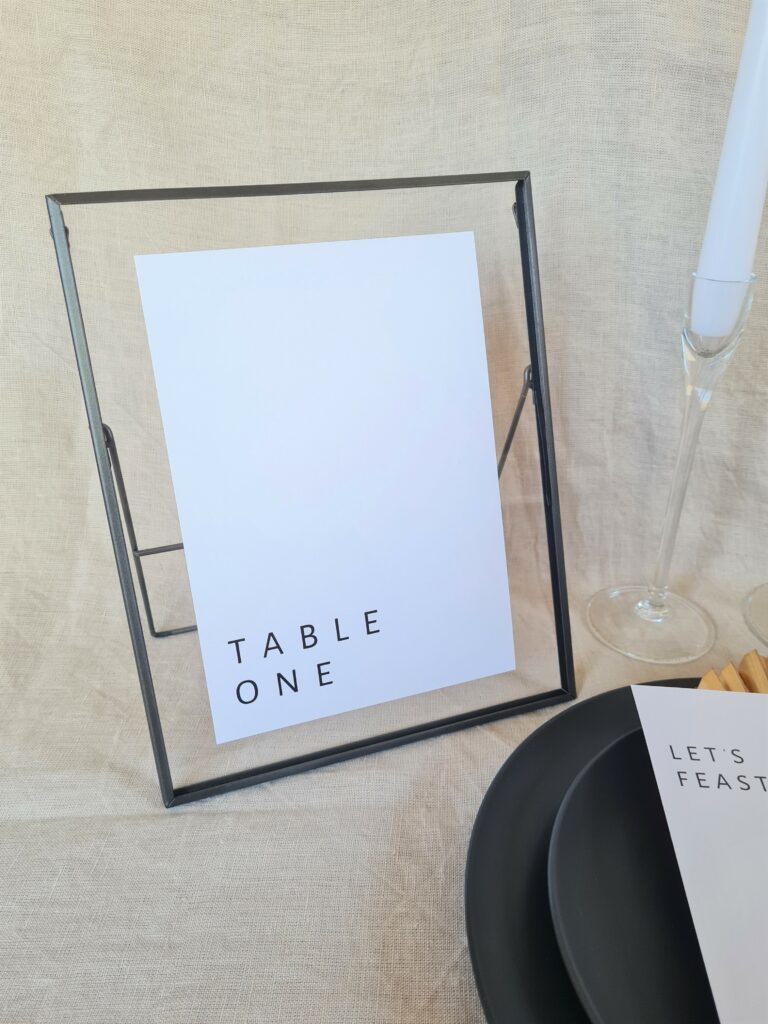 A black and white table number sign with modern text reading "Table One" placed in a black frame. The sign is placed on a table with minimal wedding décor in black, white, and gold colours.