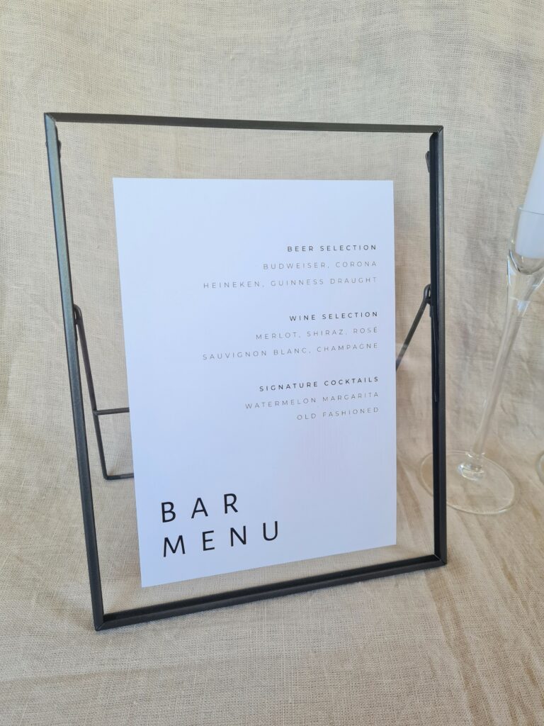 A black and white bar menu sign with modern text reading "bar menu" in a black frame. The sign is placed on a table with minimal wedding décor, including a white candle and beige linen background.