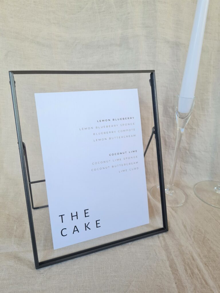 A black and white cake menu sign with modern bold text reading "the cake" in a black frame. The sign is placed on a table with minimal wedding décor, including a white candle and beige linen background.