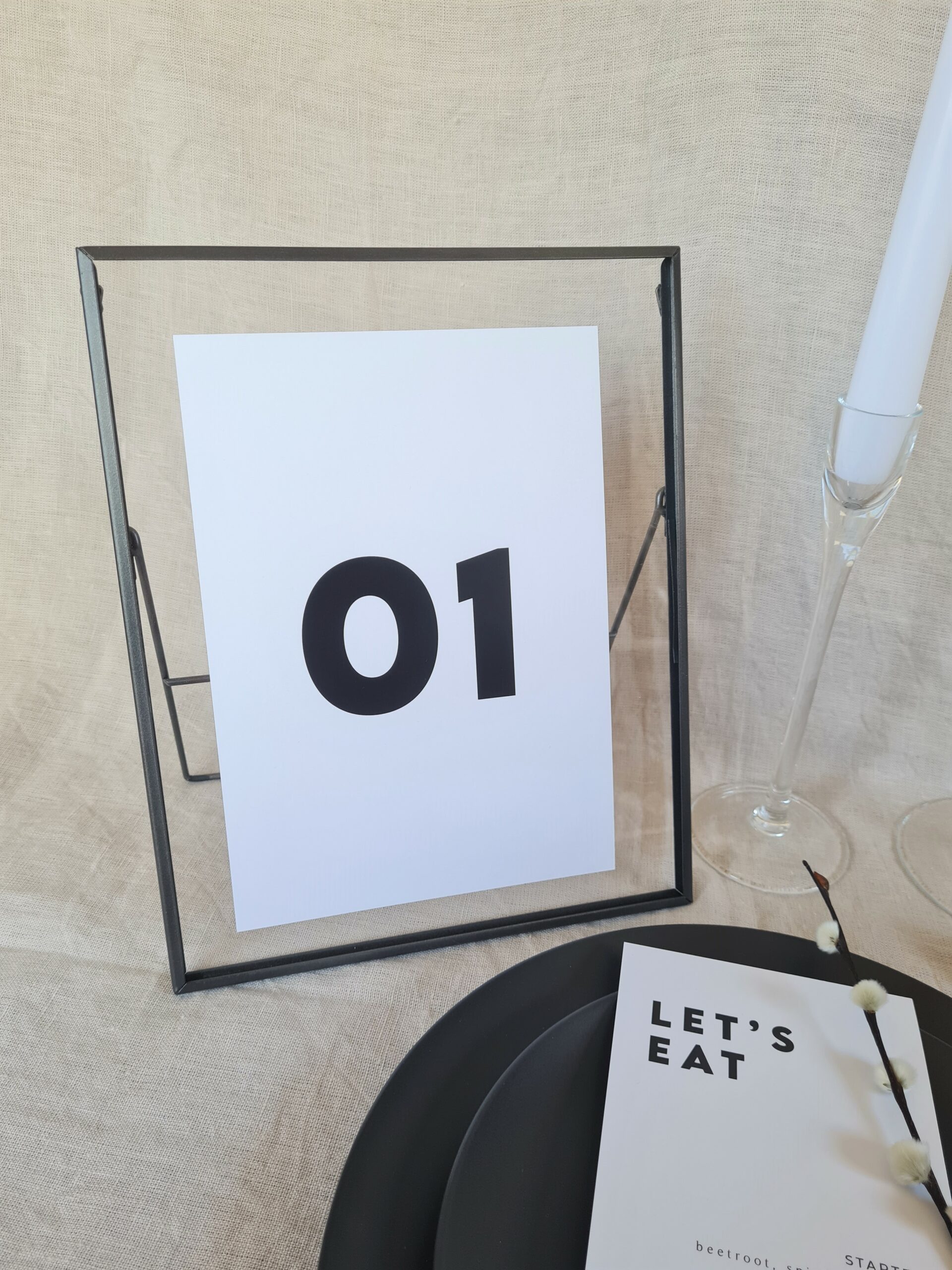 A black and white table number sign with modern bold text reading "01" placed in a black frame. The sign is placed on a table with minimal wedding décor in black, white, and nude colours