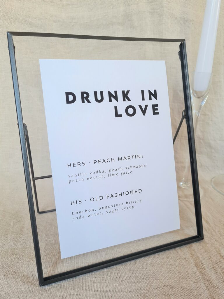 A black and white bar menu sign with modern bold text reading "drunk in love" in a black frame. The sign is placed on a table with minimal wedding décor, including a white candle and beige linen background.