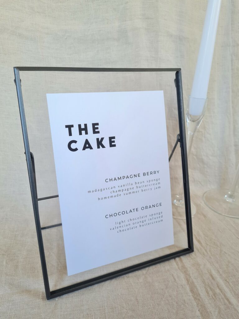 A black and white cake menu sign with modern bold text reading "the cake" in a black frame. The sign is placed on a table with minimal wedding décor, including a white candle and beige linen background.
