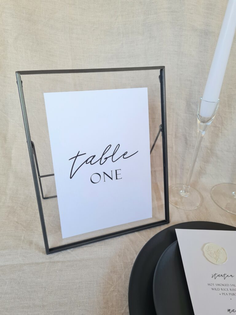A black and white table number sign with modern calligraphy-style text reading "Table One" placed in a black frame. The sign is placed on a table with minimal wedding décor in black, white, and nude colours.