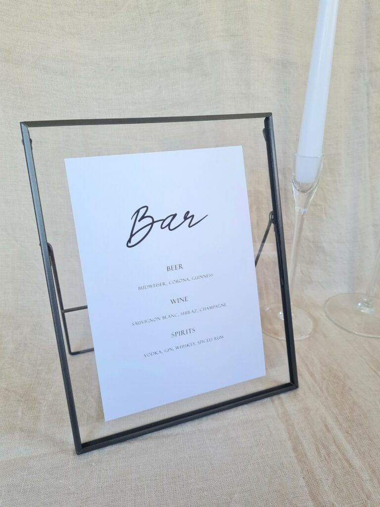 A black and white bar menu sign with modern calligraphy-style text reading "bar" in a black frame. The sign is placed on a table with minimal wedding décor, including a white candle and beige linen background.