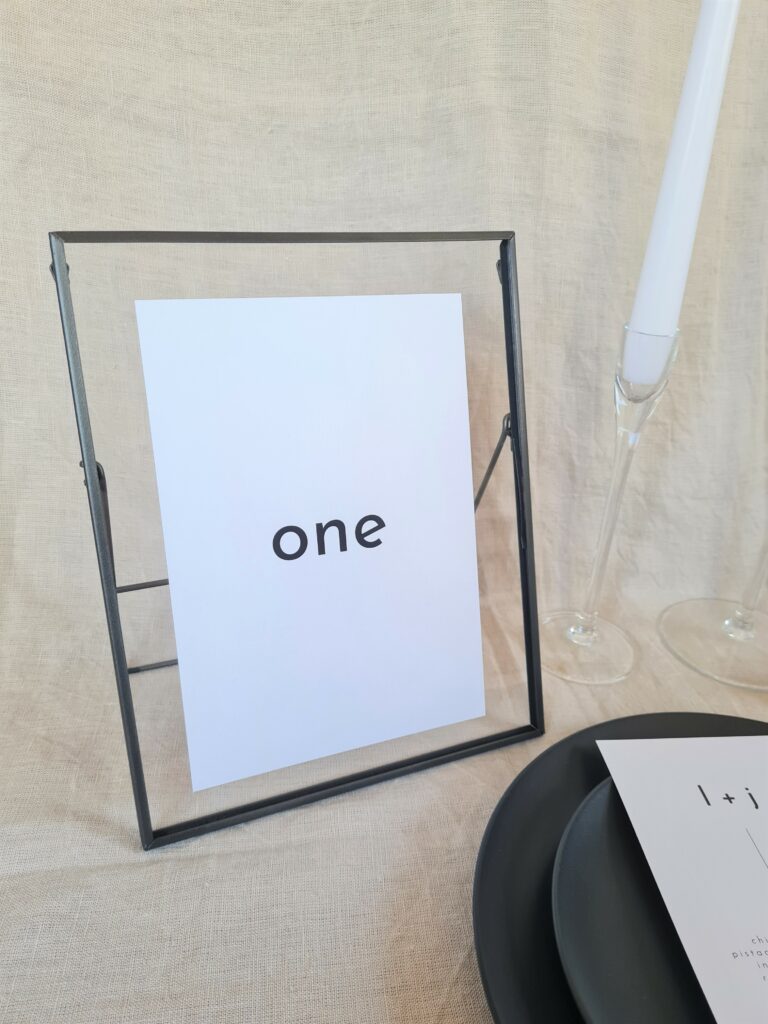 A black and white table number sign with modern bold text reading "one" placed in a black frame. The sign is placed on a table with minimal wedding décor in black, white, and nude colours