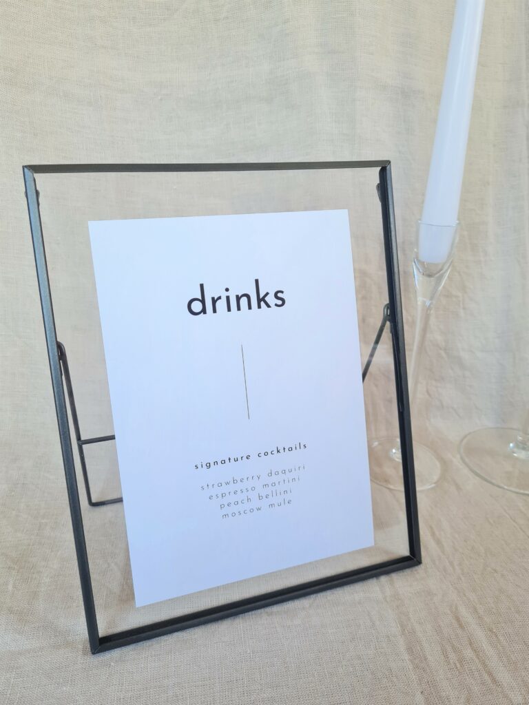 A black and white bar menu sign with modern bold text reading "drinks" in a black frame. The sign is placed on a table with minimal wedding décor, including a white candle and beige linen background.