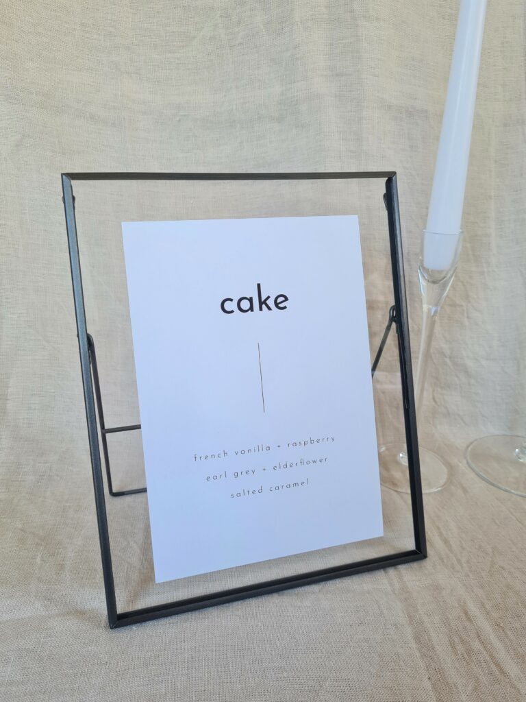 A black and white cake menu sign with modern bold text reading "cake" in a black frame. The sign is placed on a table with minimal wedding décor, including a white candle and beige linen background.