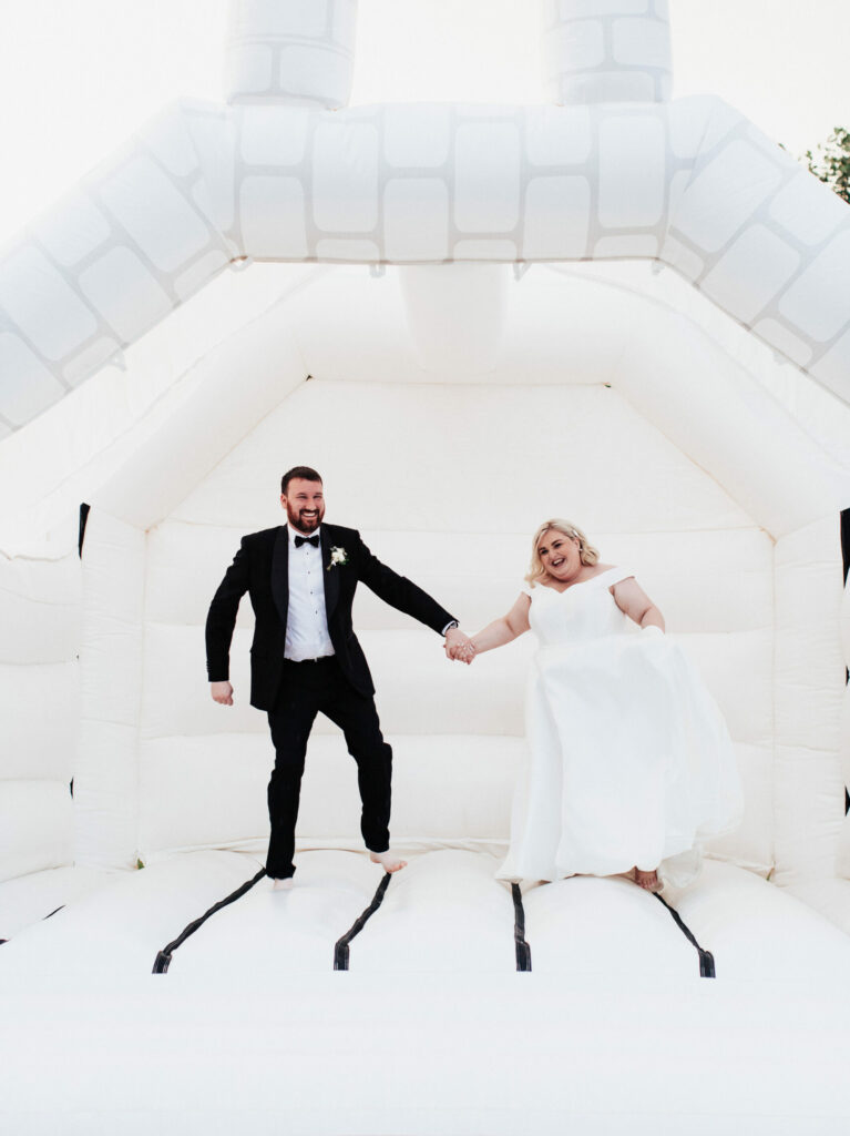 An image of a bride wearing a white wedding dress and standing next to the groom in a black tie suit. They are joyfully jumping on a white bouncy castle in the gardens of Brickhouse Vineyard near Exeter, Devon.