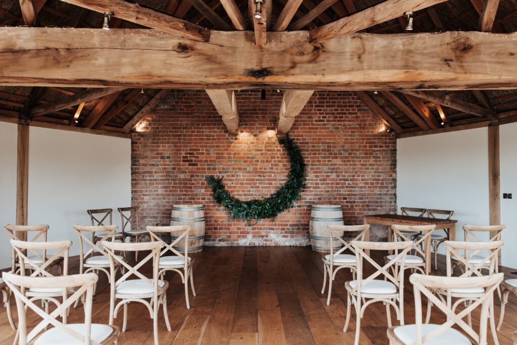 An image of the roundhouse barn wedding venue at Brickhouse Vineyard near Exeter, Devon. The venue is set up with chairs laid out in neutral tones, creating an elegant and inviting atmosphere. At the top of the aisle, there is a beautiful green foliage crescent moon installation, adding a touch of natural and whimsical charm to the setting.