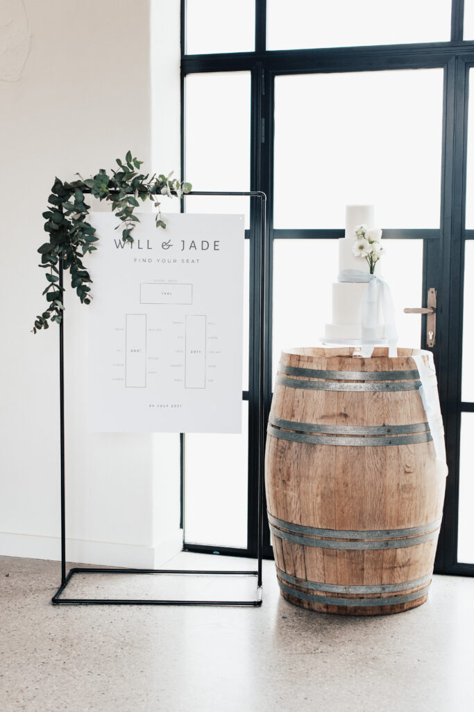 The image features the beautiful inside of the barn at Brickhouse Vineyard located near Exeter, Devon. The picture showcases the a black frame sign with a bespoke hanging seating plan sign reading " Will & Jade, find your seat". It is beautifully decorated with green foliage and is set next to a wooden wine barrel with a white wedding cake sat on top elegantly decorated with white flowers and a blue ribbon.