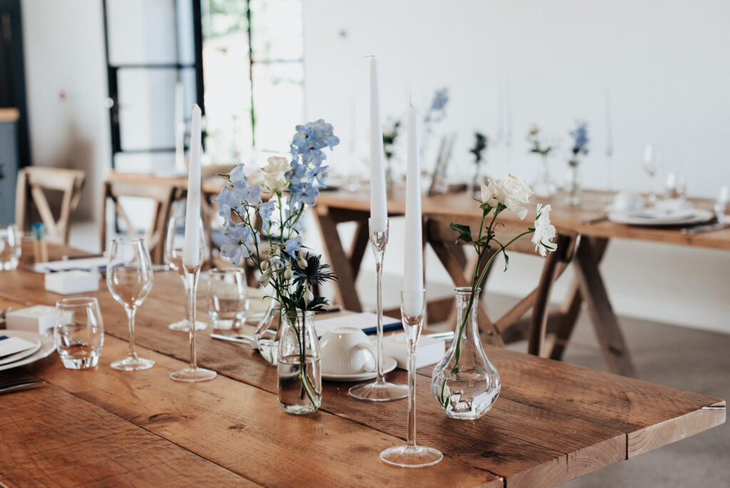 An image inside the wedding barn at Brickhouse Vineyard near Exeter, Devon. The photo showcases the rustic charm of the venue, with trestle tables set up for the wedding reception. The tables are adorned with natural-toned decor, including hints of blue flowers, adding a touch of color and elegance to the space. The combination of the rustic elements and delicate floral accents creates a warm and inviting atmosphere.