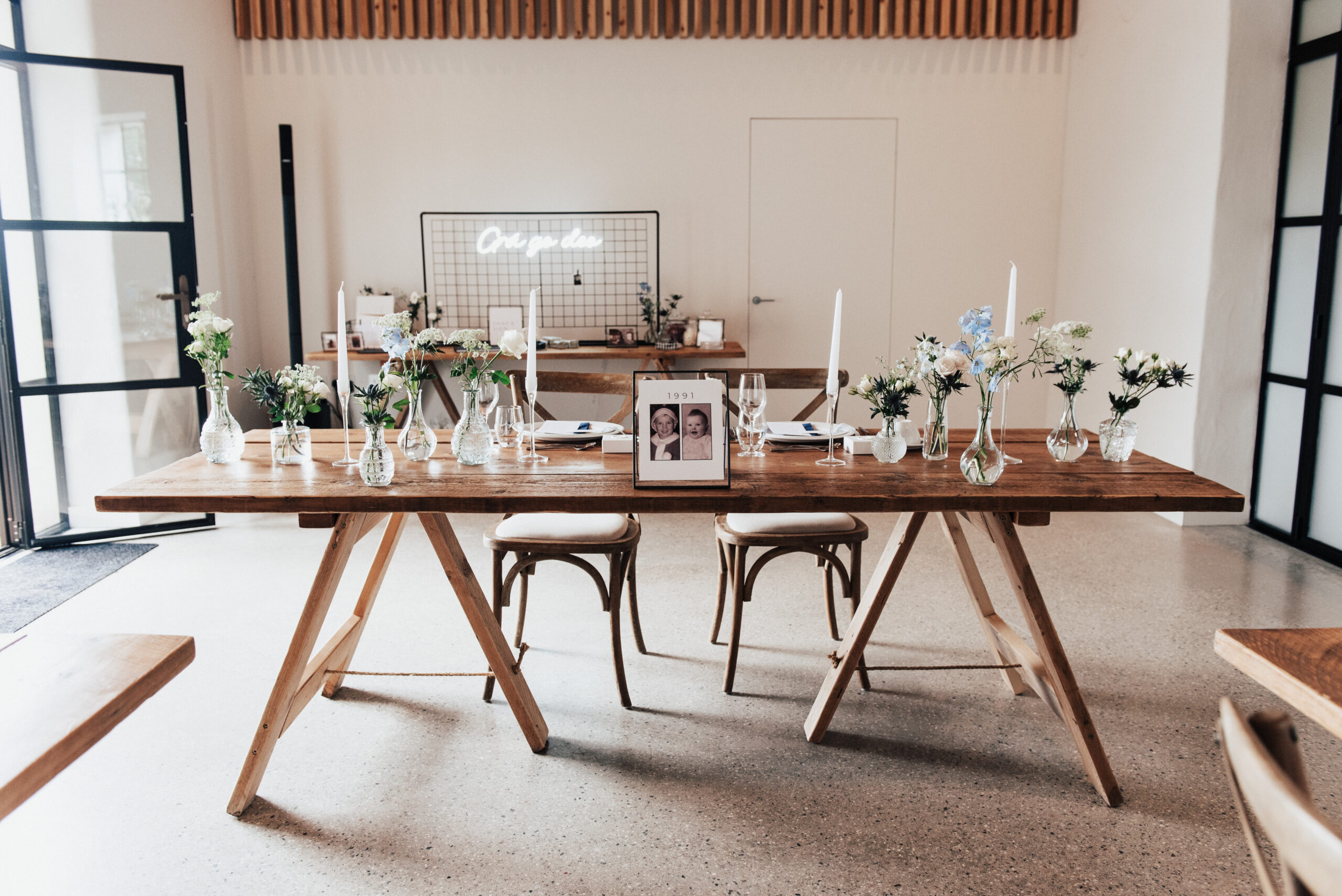 A black and white table name sign that reads "1991" with childhood photos of the bride and groom is displayed in a black metal frame on a wooden table. White candles and blue flowers are placed next to the menu. There is a neon sign in the background with a black frame. This setting is at a wedding at Brickhouse Vineyard in Devon.