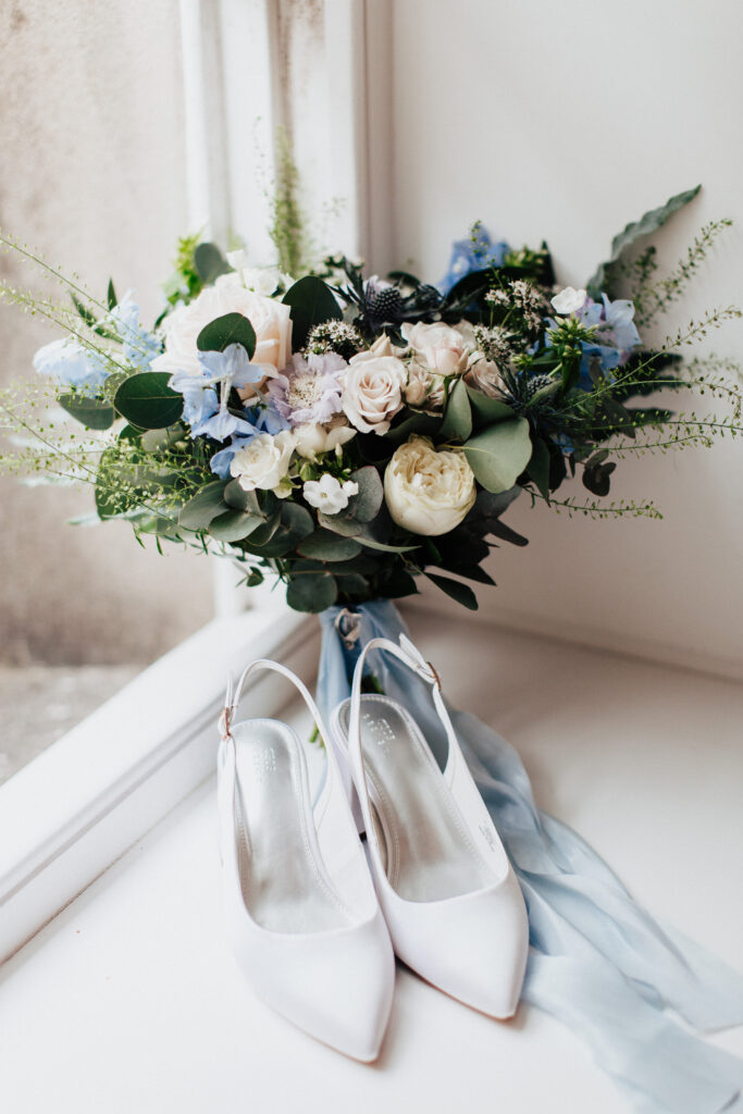 An image of white silk wedding heels placed next to a stunning modern bouquet. The bouquet is crafted with a combination of white, cream, and blue flowers, along with lush greenery. It is beautifully tied together with a blue silk ribbon, adorned with an antique silver Irish harp pin. The elegant and delicate details of the bouquet and the stylish white silk heels create a sense of sophistication and bridal charm.