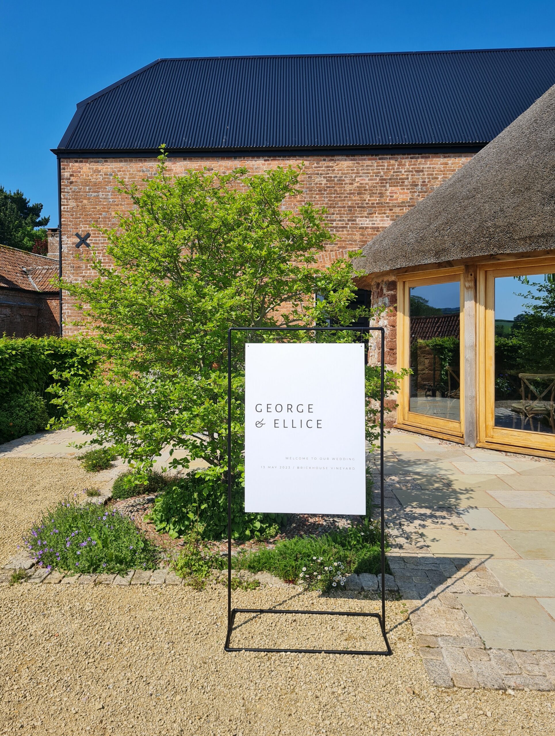 A black and white minimal and modern wedding welcome sign placed in front of the stunning red brick barn and thatched roundhouse at Brickhouse Vineyard in Exeter, Devon. The sign is made of matte black copper and features sleek and contemporary typography. The contrast between the black sign and the red brick barn creates a visually striking image. The surrounding gardens add a touch of natural beauty to the scene.