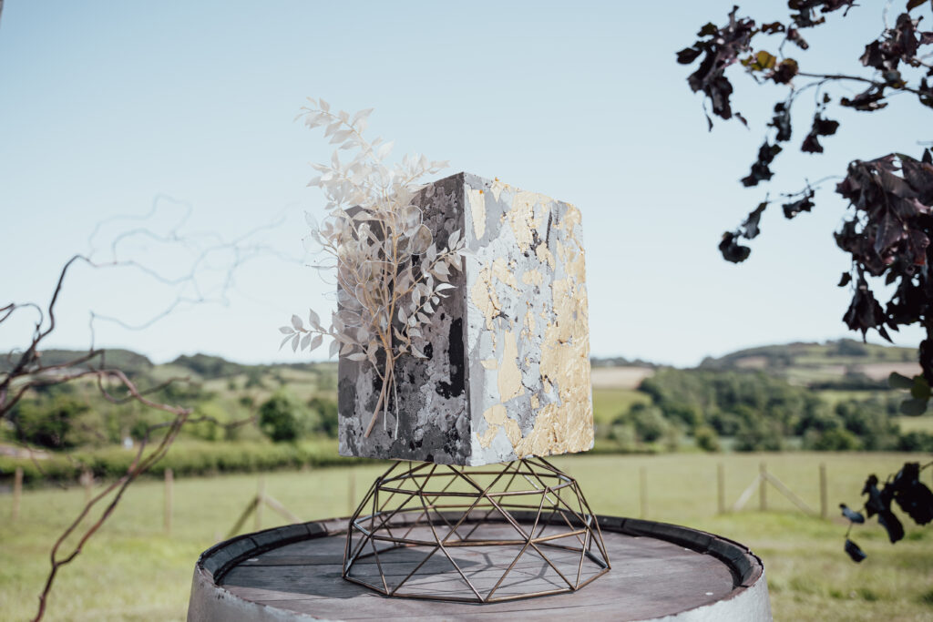 A stunning grey and white marbled wedding cake sits on a dome-wired cake stand. The cake is adorned with delicate white dried flowers and intricate gold leaf detailing. In the background, picturesque countryside views add a touch of natural beauty to the scene, creating a picturesque setting for the wedding celebration.